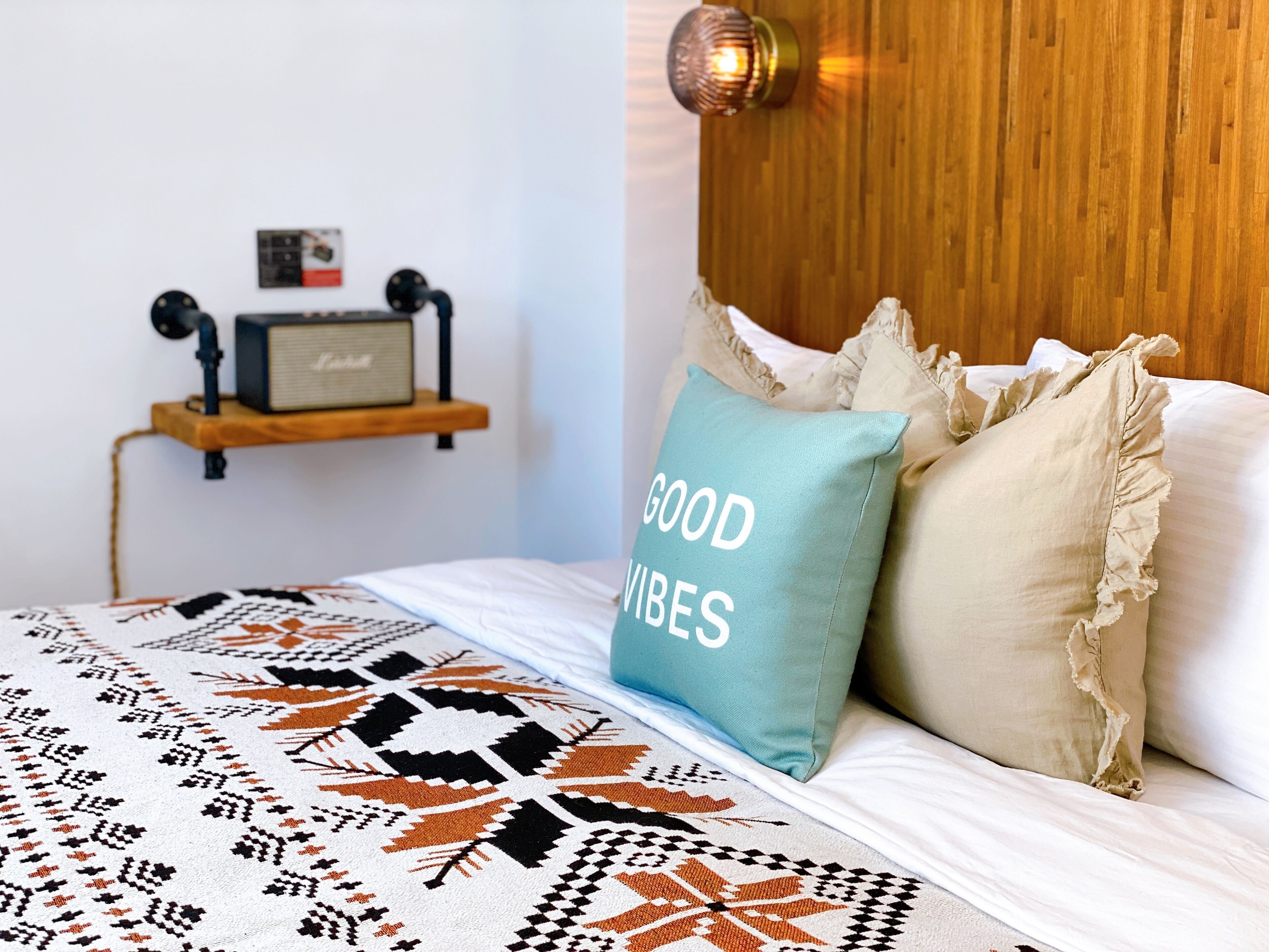 A bedroom with a bed, a wall-mounted shelf with a radio, and a pillow with "GOOD VIBES" text.