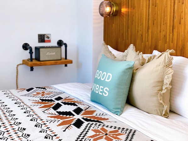 A bedroom with a bed, a wall-mounted shelf with a radio, and a pillow with "GOOD VIBES" text.