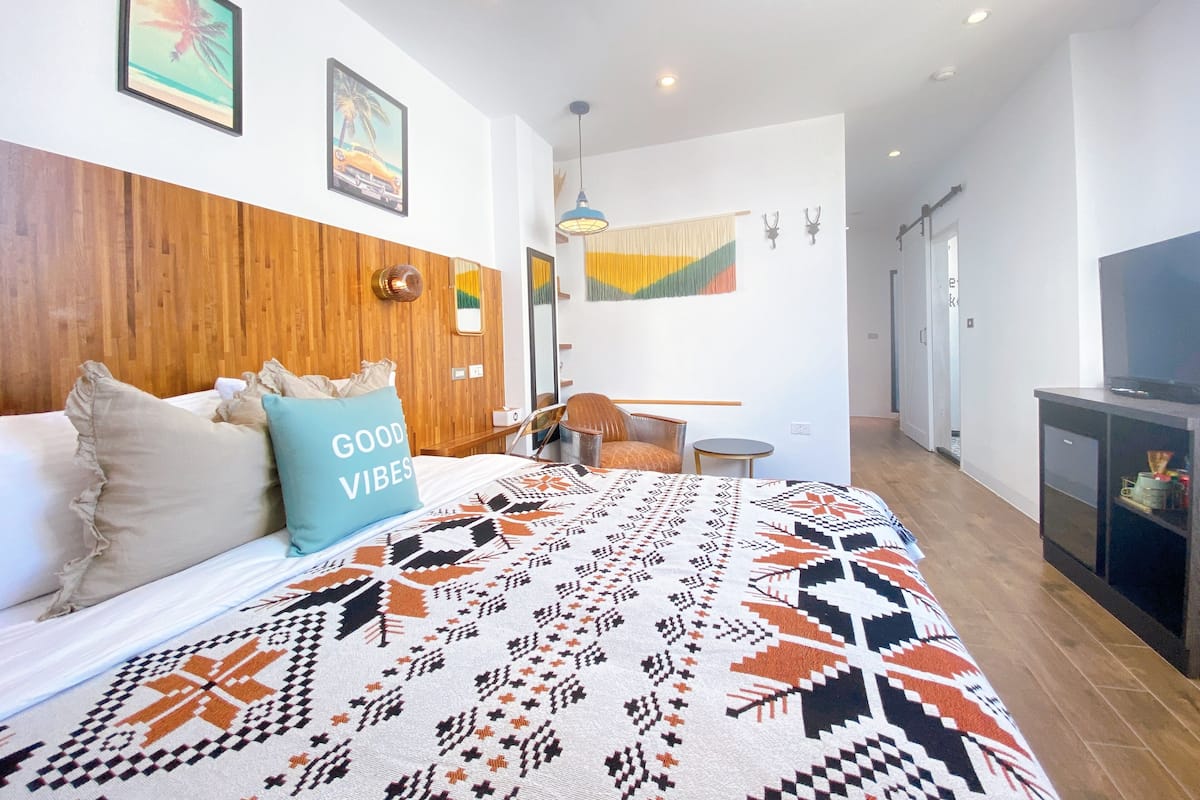 A modern bedroom with a wooden headboard, a patterned bedspread, and a "GOOD VIBES" pillow.