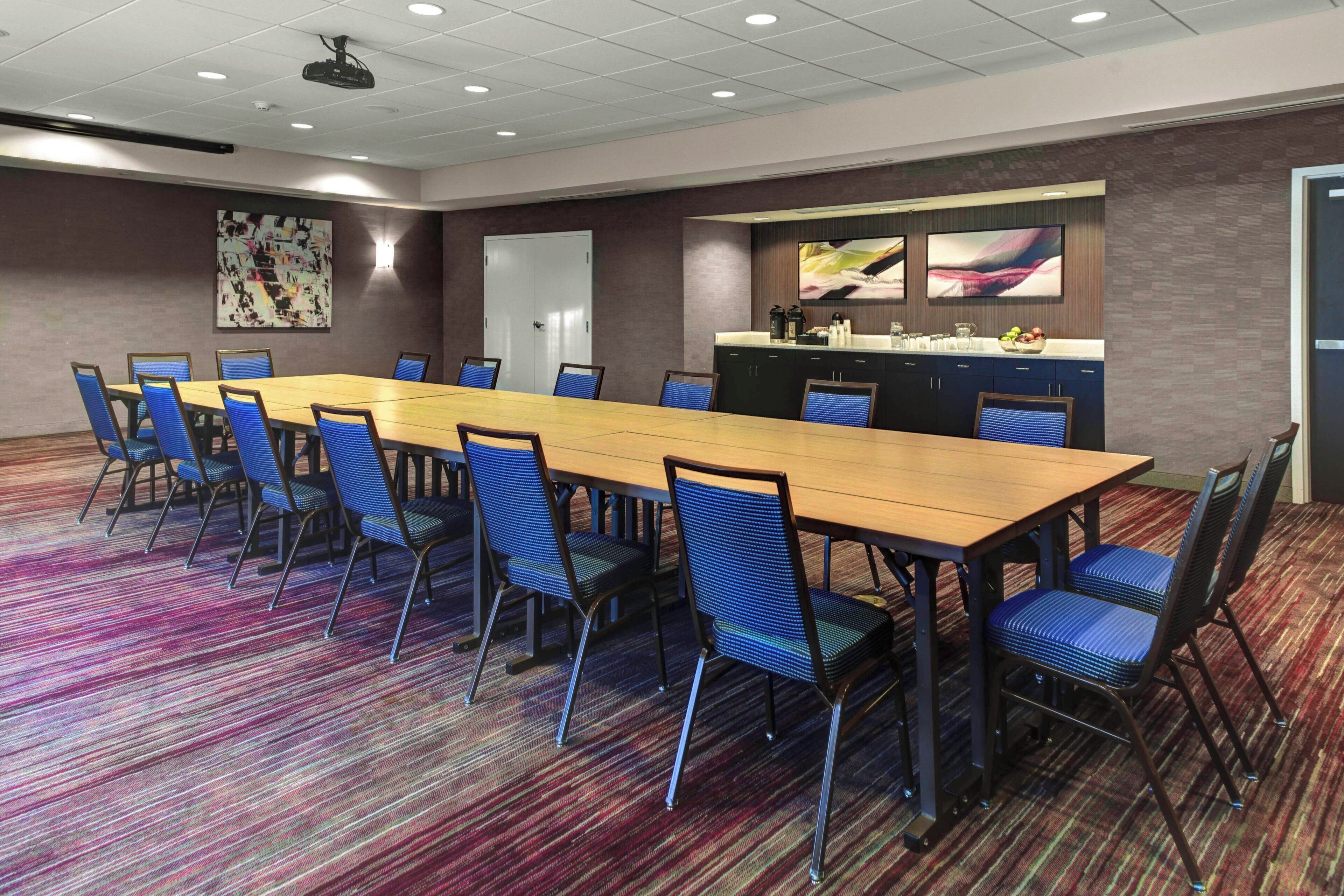 Un salón de conferencias con una mesa larga, sillas azules, una zona de cocina y una puerta.