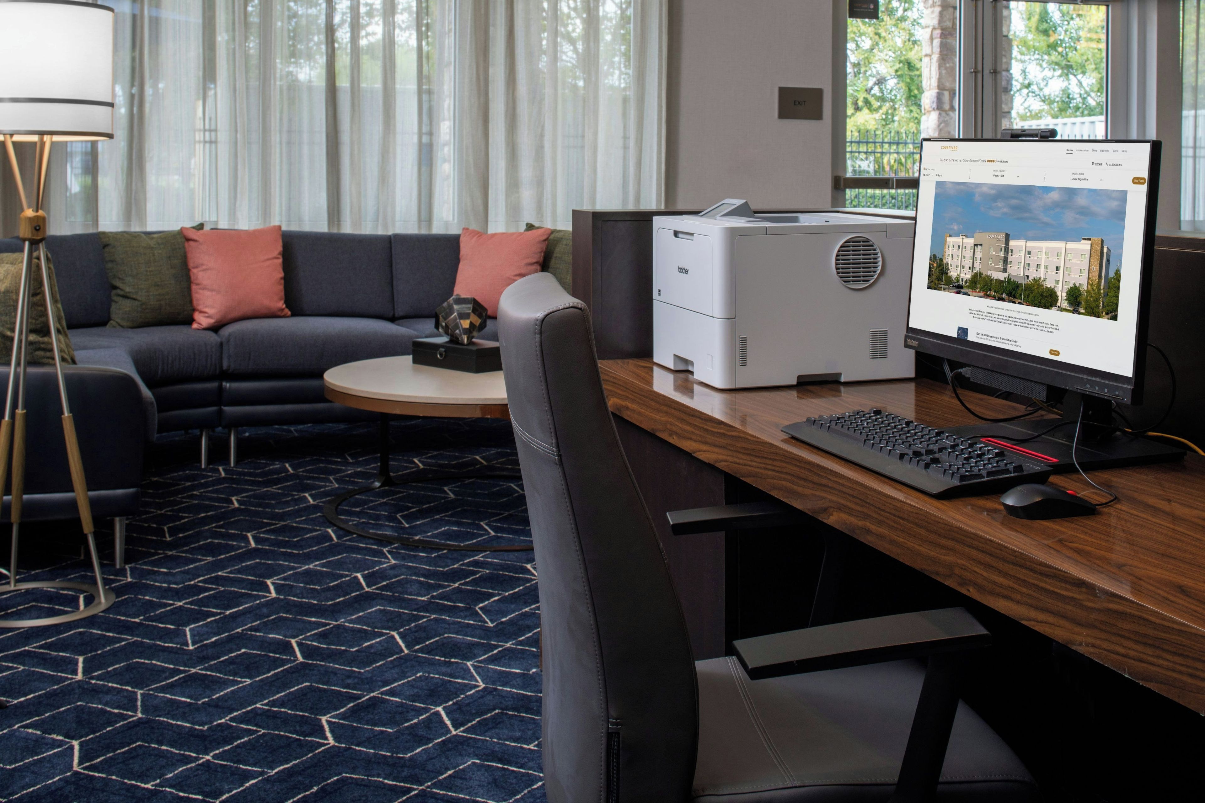 A modern hotel room with a dark blue carpet, a sofa with colorful cushions, a wooden desk with a computer, and a printer.