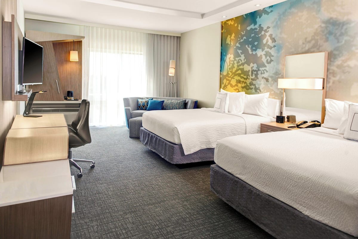 Une chambre d’hôtel avec deux lits, un bureau, une chaise, une télévision et une grande fenêtre avec des rideaux.