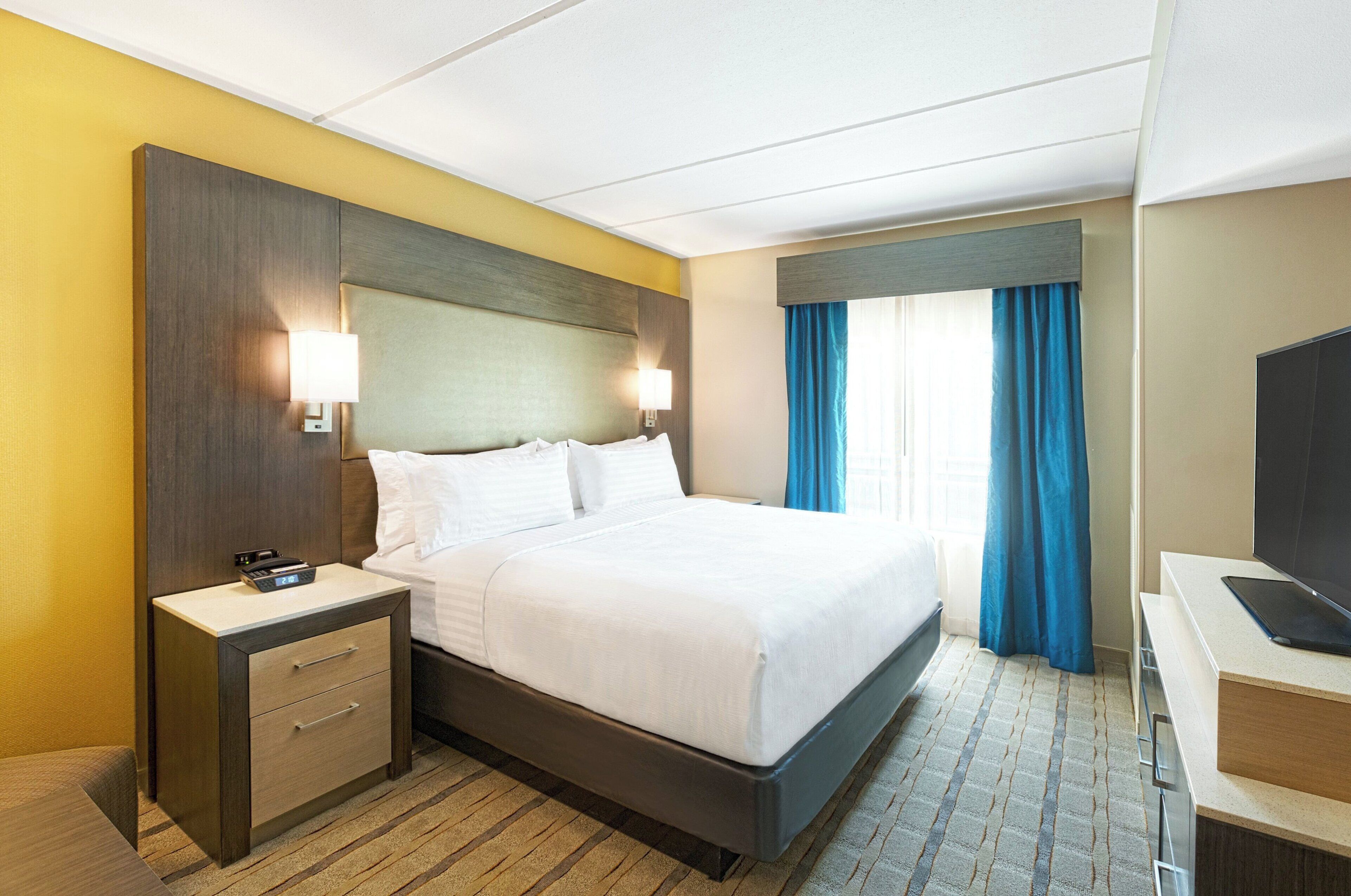 Quarto de hotel com cama, mesas de cabeceira, escrivaninha com televisão de tela plana e janela com cortinas azuis.
