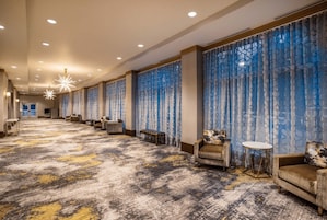 Pasillo de hotel con alfombra estampada, zonas de estar y cortinas azules con diseño.
