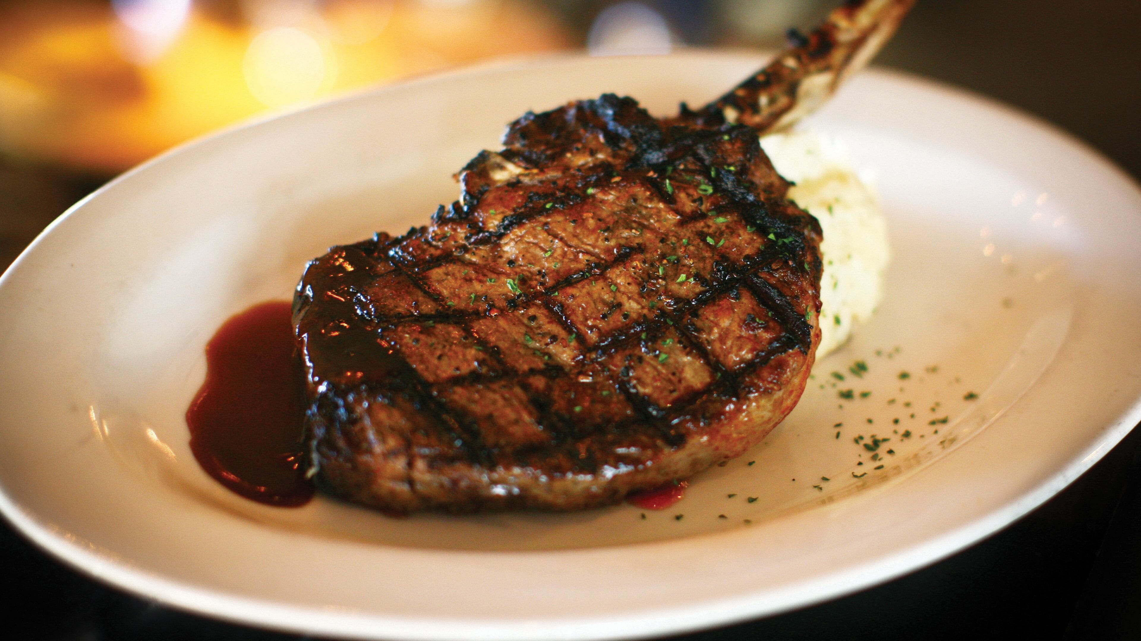 A steak with a charred exterior, served on a white plate with a side of mashed potatoes and a red sauce.
