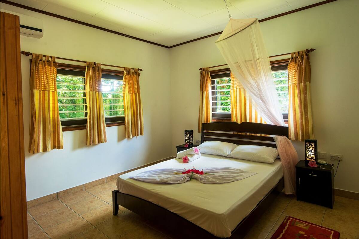 A neatly made bed with a white bedspread, a decorative pillow, and a bouquet of flowers. There are two bedside tables with decorative items and a nightstand with a lamp. The room has a wooden headboard and curtains on the windows.