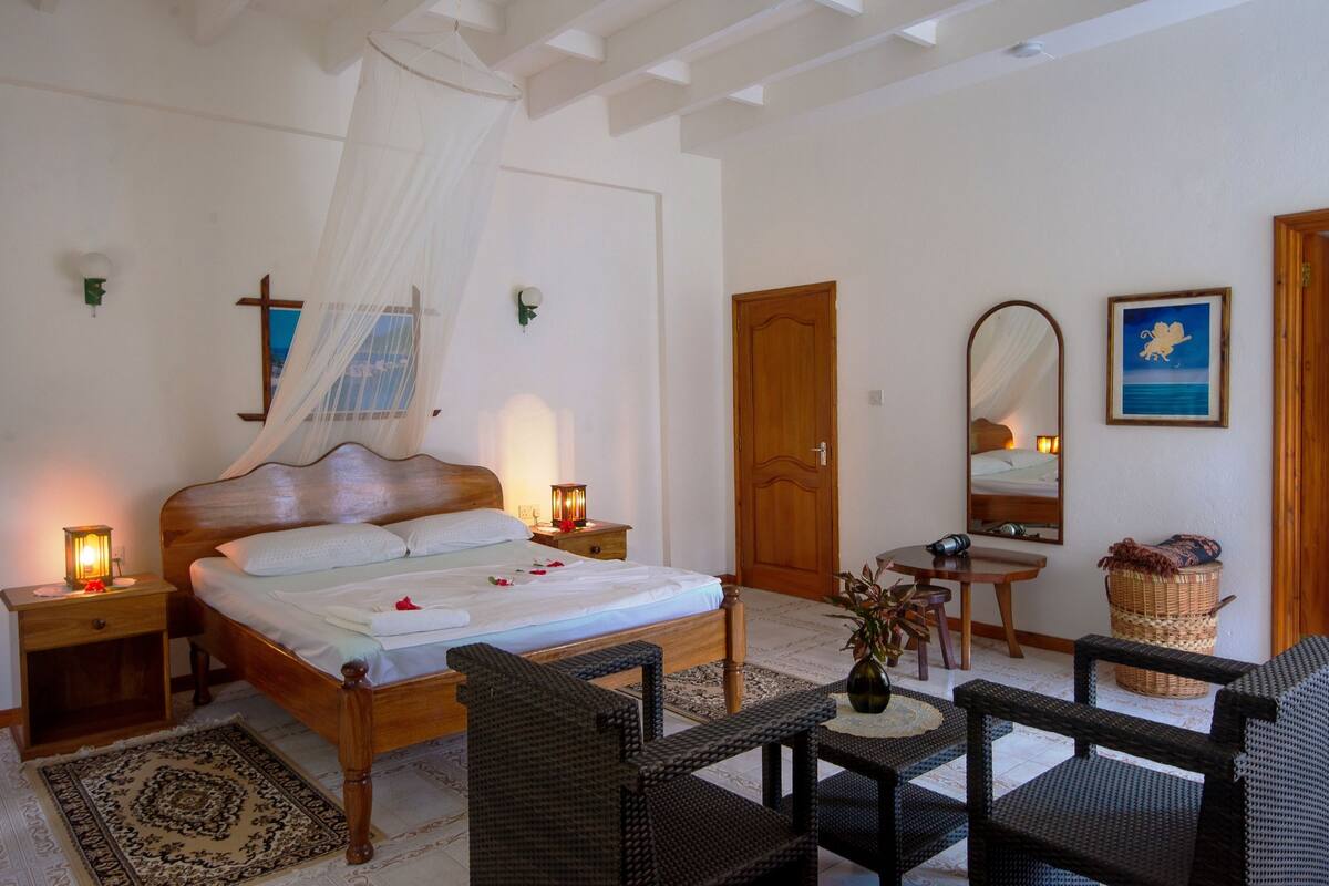 A bedroom with a wooden bed, bedside lamps, a nightstand, a wicker chair, a small table, a mirror, and a door.