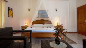 Quarto com cama sob um dossel branco, duas luminárias de cabeceira, uma cadeira de vime, uma mesinha de cabeceira e um vaso com planta.