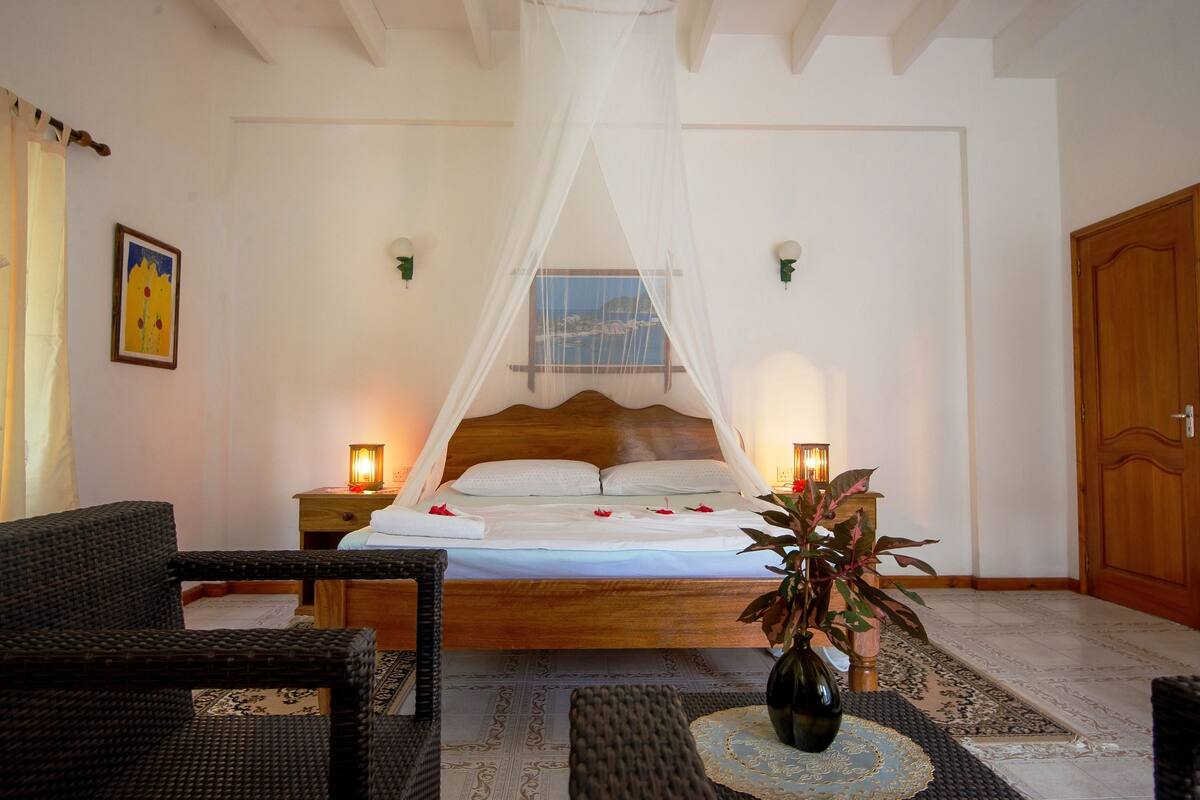 A bedroom with a bed under a white canopy, two bedside lamps, a wicker chair, a bedside table, and a potted plant.