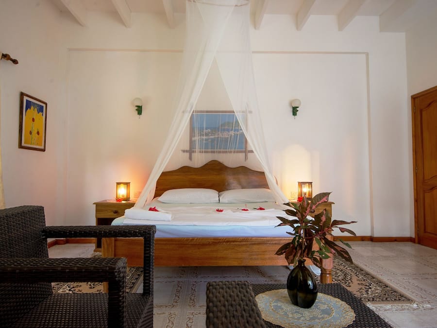 Un dormitorio con cama bajo dosel blanco, dos lámparas de noche, una silla de mimbre, una mesita de noche y una planta en maceta.