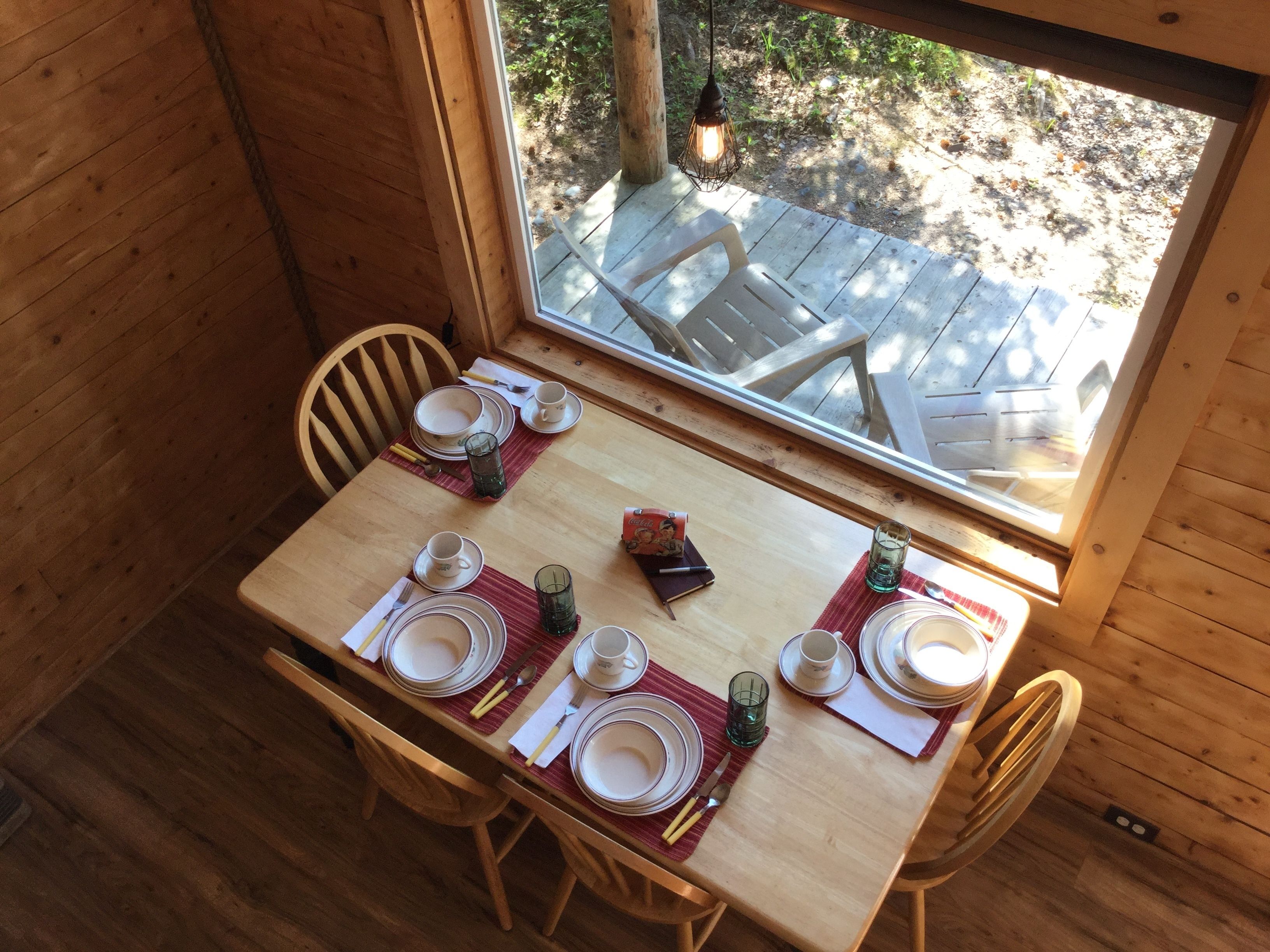 A wooden dining table set with plates, glasses, and cutlery, with a view of an outdoor deck through a window.