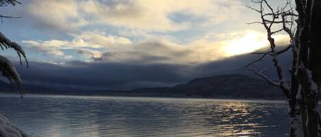 A serene lakeside view with snow-covered ground and bare trees, under a cloudy sky.