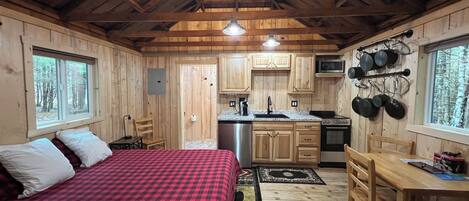 A cabin interior with a bed, wooden walls, a kitchen area, and a dining table.