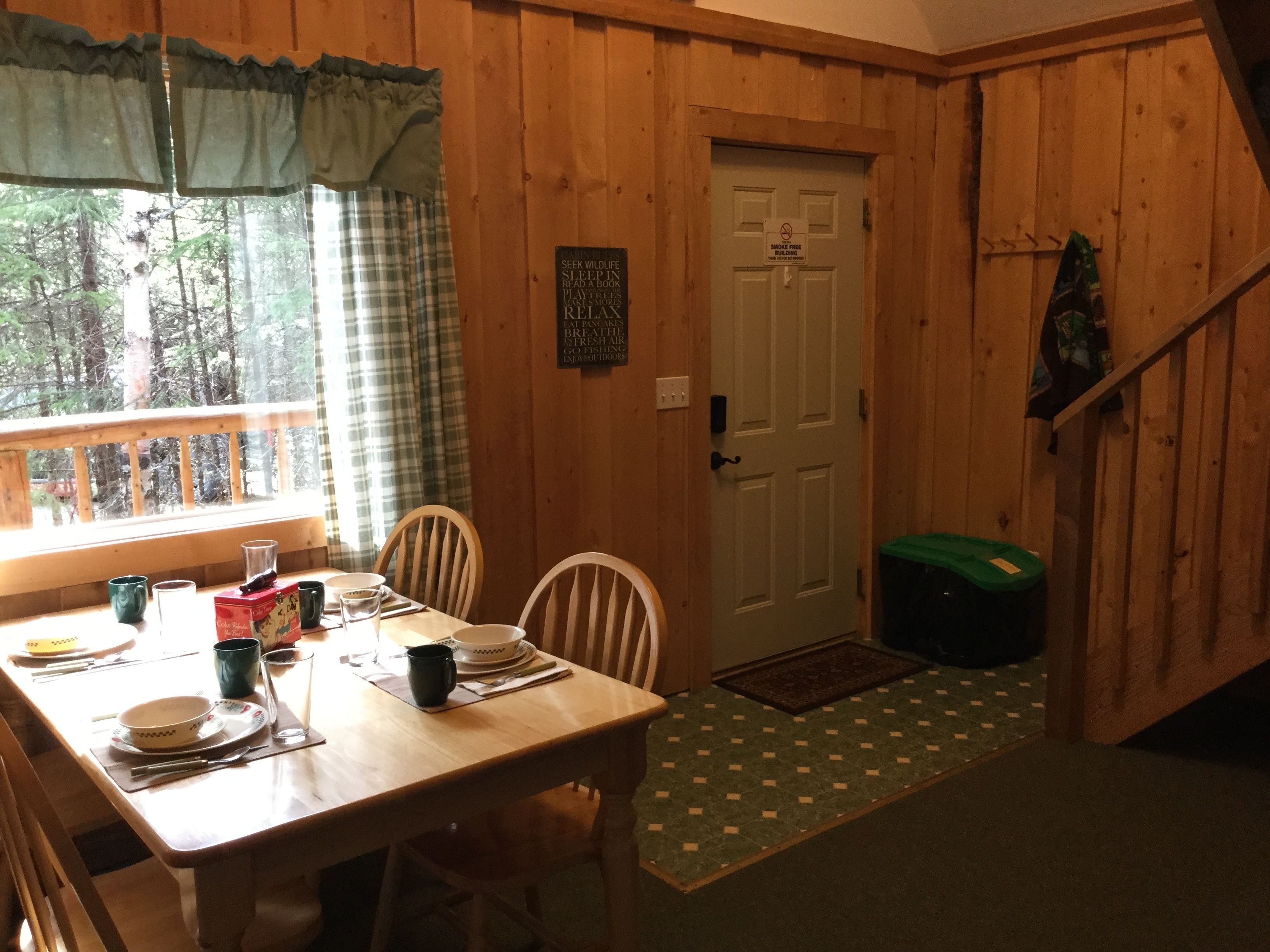 A wooden cabin interior with a dining table set for four, a window with curtains, a door with a 'No Smoking' sign, and a green trash bin.