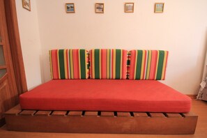 Una cama con colchón rojo y almohadas a rayas, un banco de madera al pie y cuadros en la pared.