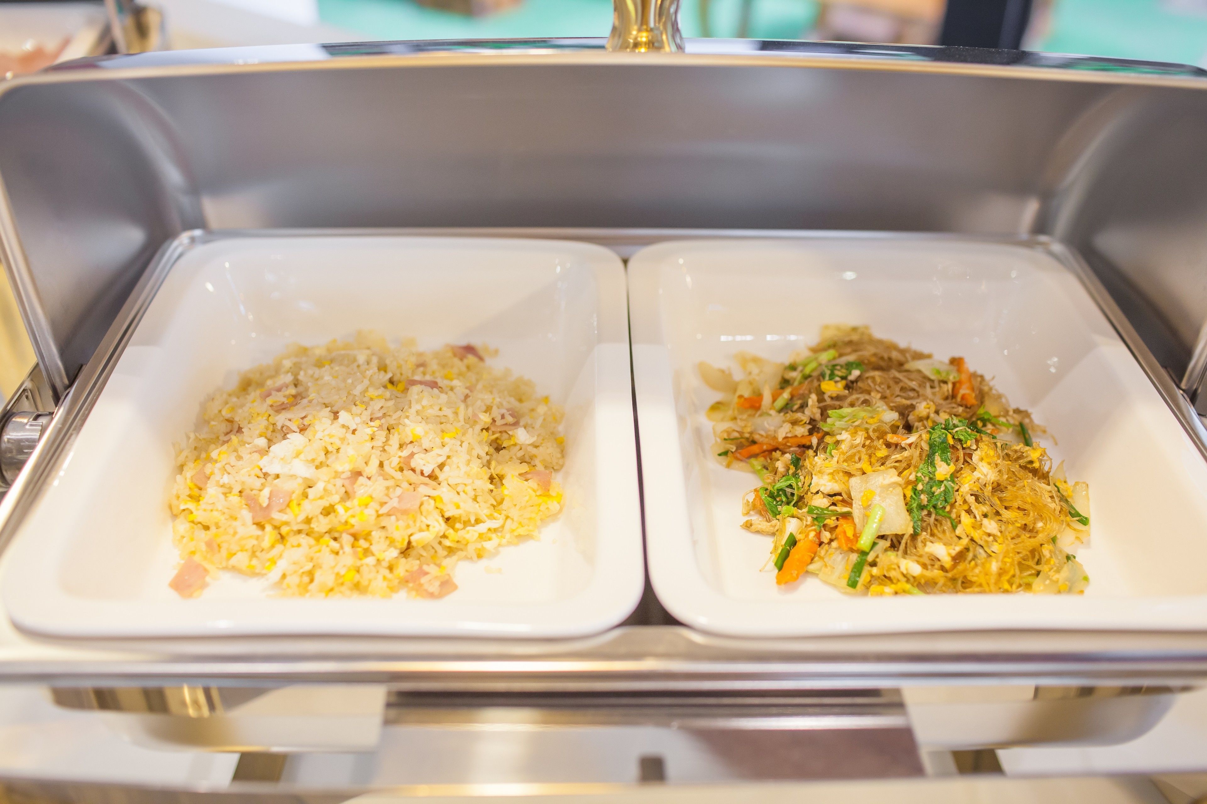 Two white rectangular dishes of fried rice, one with visible ham and the other with vegetables, in a stainless steel buffet setting.