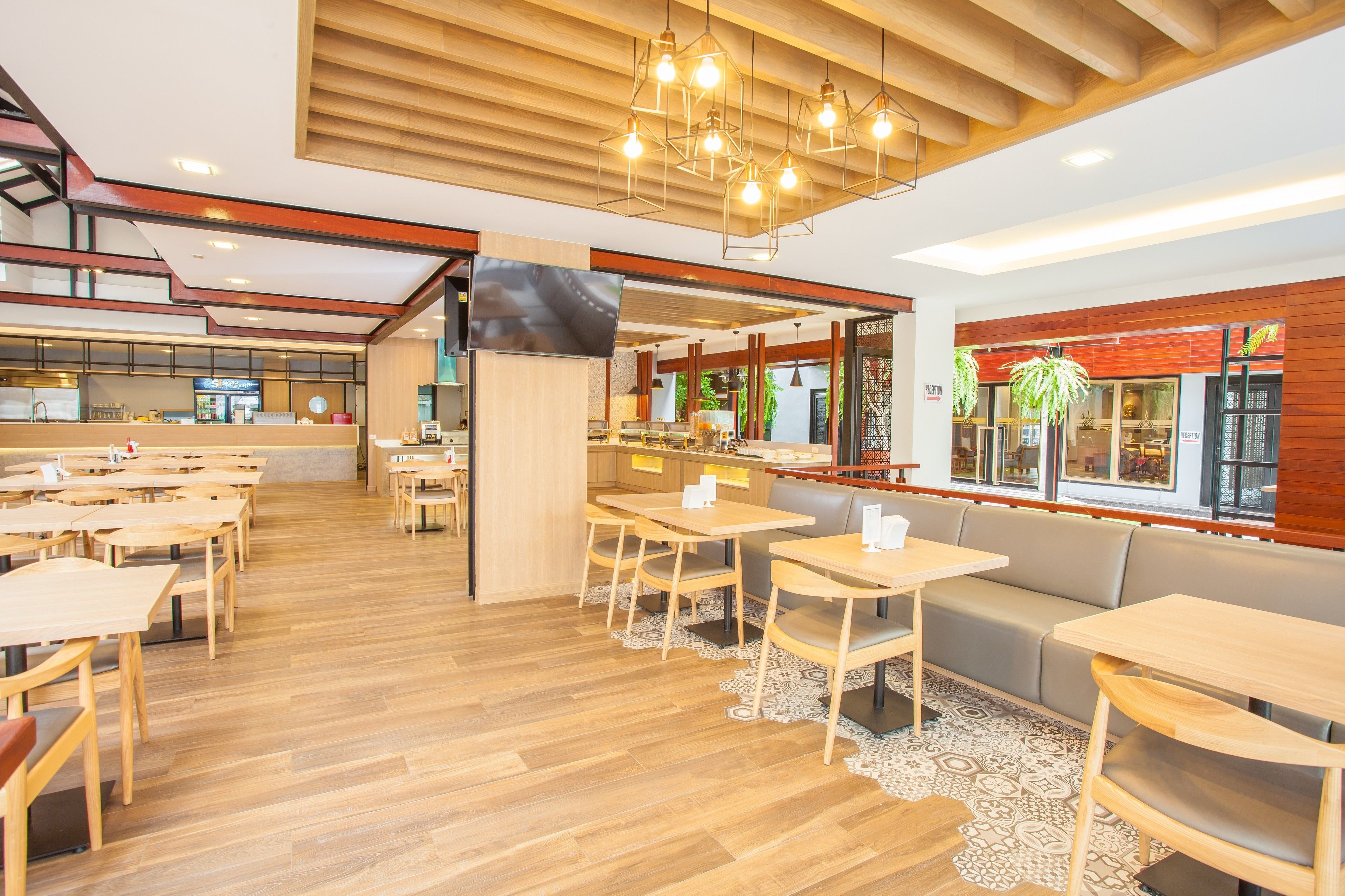 Un restaurante espacioso con suelo de madera, varias zonas de estar con mesas y un sofá, una pantalla de televisión y grandes ventanales con luz natural.