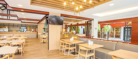 Un restaurante espacioso con suelo de madera, varias zonas de estar con mesas y un sofá, una pantalla de televisión y grandes ventanales con luz natural.