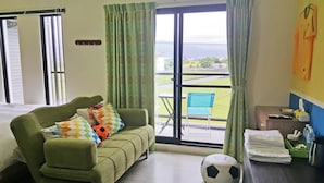 Habitación de hotel con sofá verde, cama, balcón con vistas, pelota de fútbol y mesa con toallas y artículos de aseo.