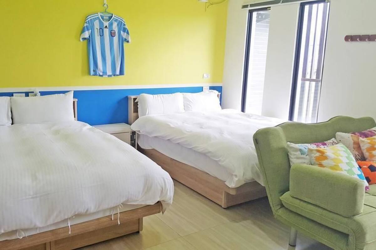 A room with two beds, a green armchair, and a window with blinds.