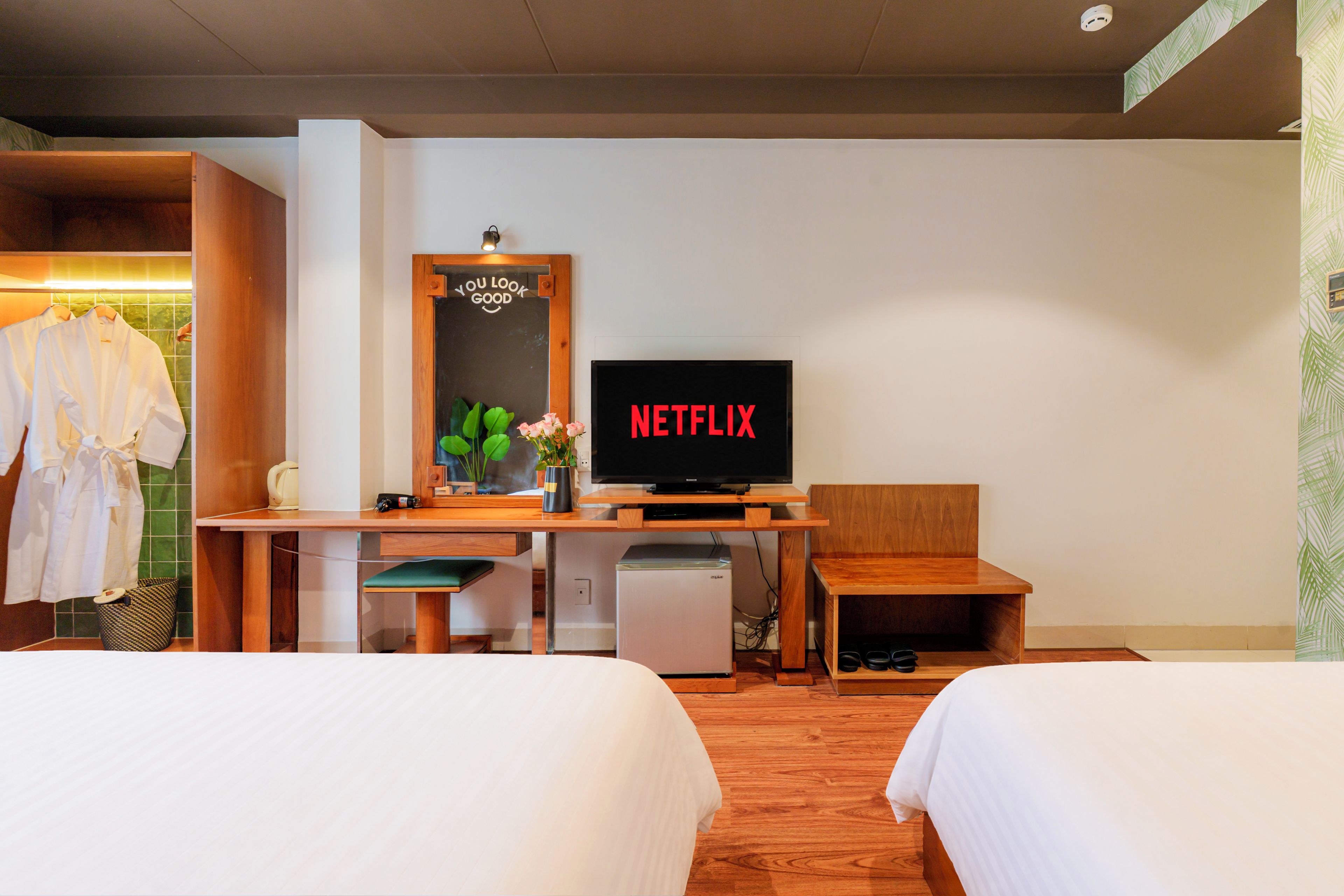 Ein Hotelzimmer mit zwei Betten, einem Schreibtisch mit Fernseher, der 'NETFLIX' anzeigt, einer kleinen Kühlschrank und einem Holzstuhl.