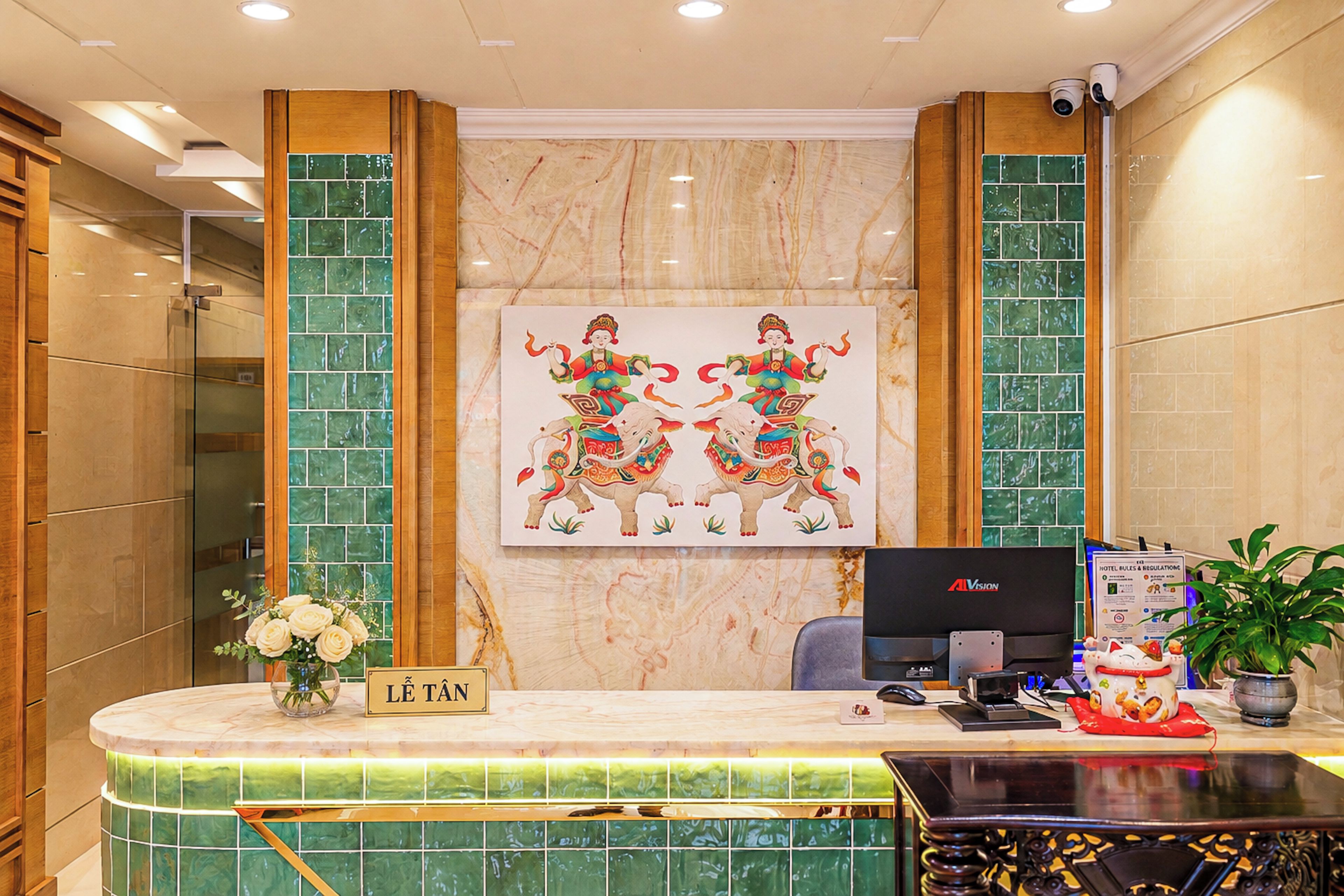 Una recepción de hotel con una computadora, un cartel que dice 'Lê Tân' y una pared decorativa con una pintura.