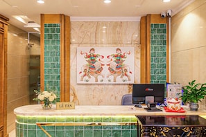 Un comptoir d’accueil d’hôtel avec un ordinateur, une enseigne indiquant « Lê Tân », et un mur décoratif orné d’un tableau.