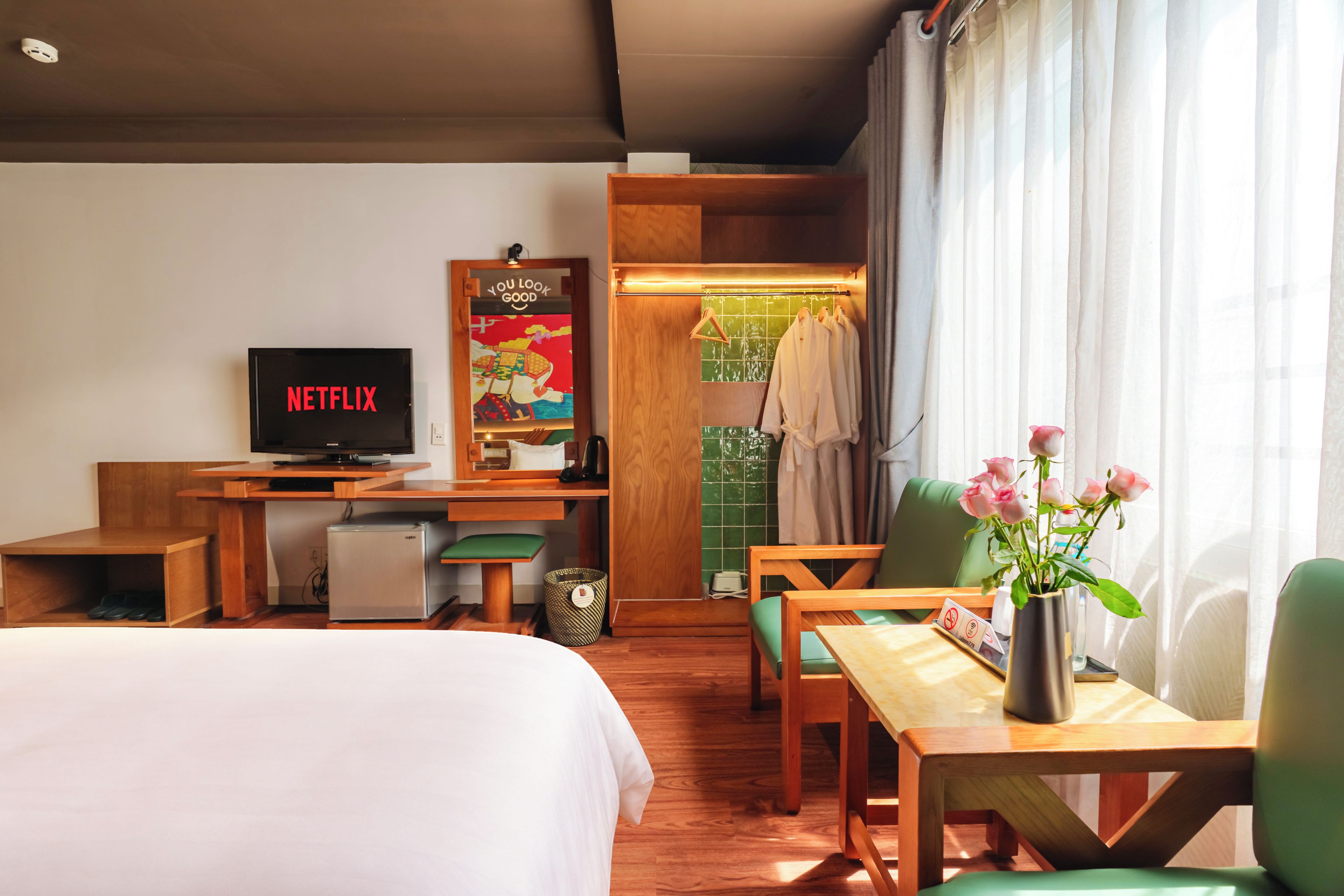 Une chambre d’hôtel avec un lit, un bureau sur lequel est posée une télévision diffusant du contenu « NETFLIX », un petit réfrigérateur, une chaise verte, une table en bois et un vase avec des fleurs.