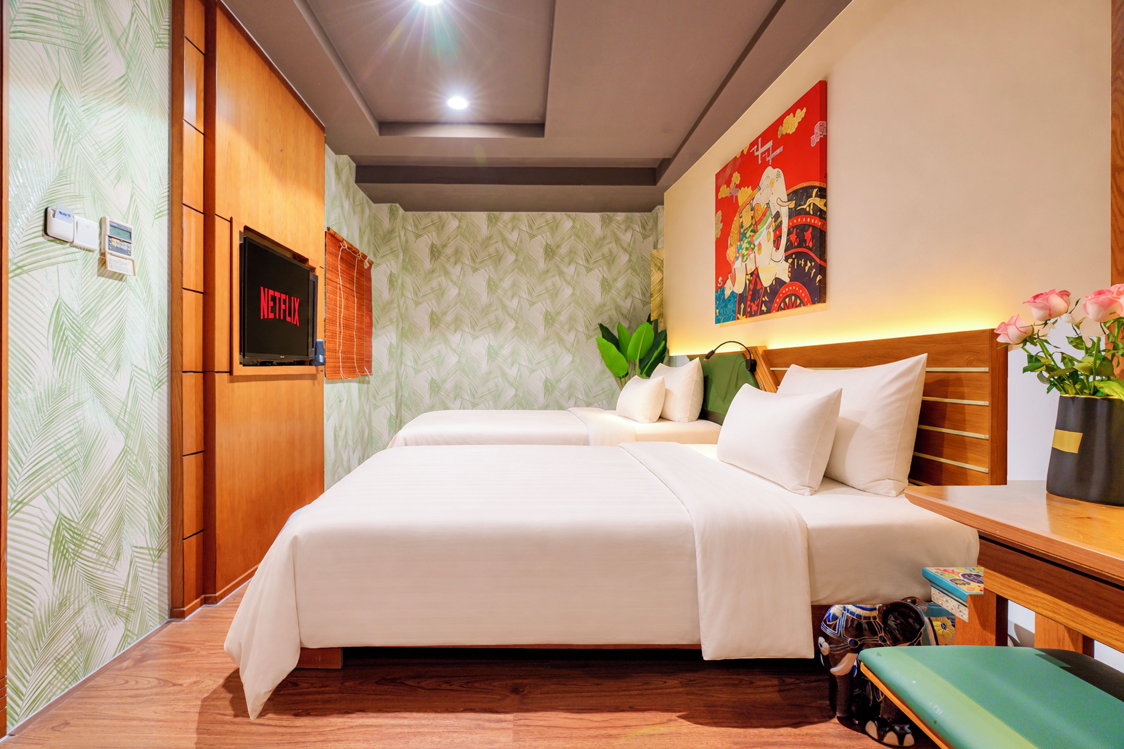 A hotel room with a bed, a television, a wall-mounted thermostat, a painting, and a plant.