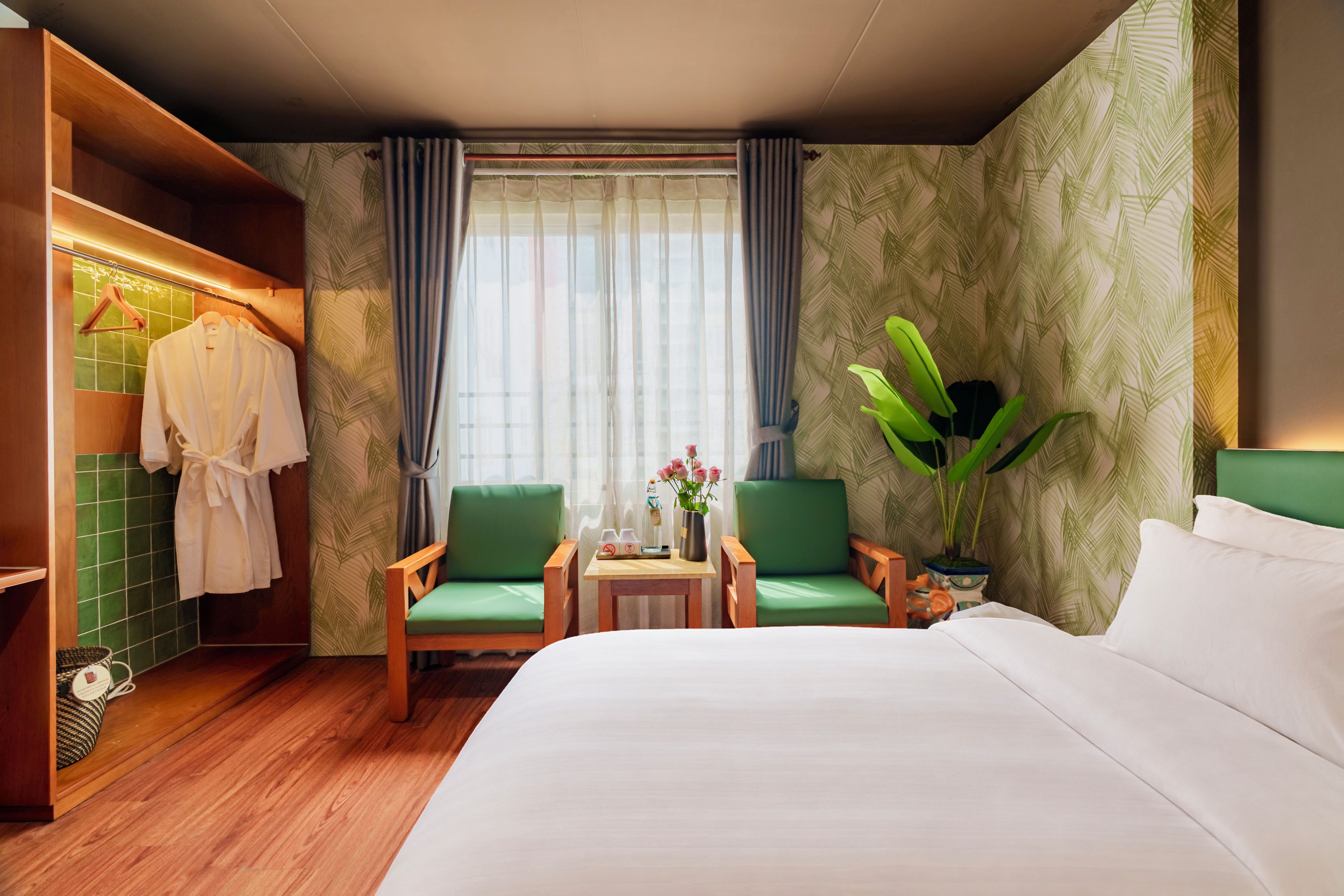 A hotel room with a bed, two chairs, a small table with a vase of flowers, a coat rack with robes, and a large potted plant.