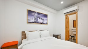 Ein Hotelzimmer mit Bett, Bank, einem Bild einer Brücke, einer Klimaanlage und einem sichtbaren Badezimmer durch eine offene Tür.