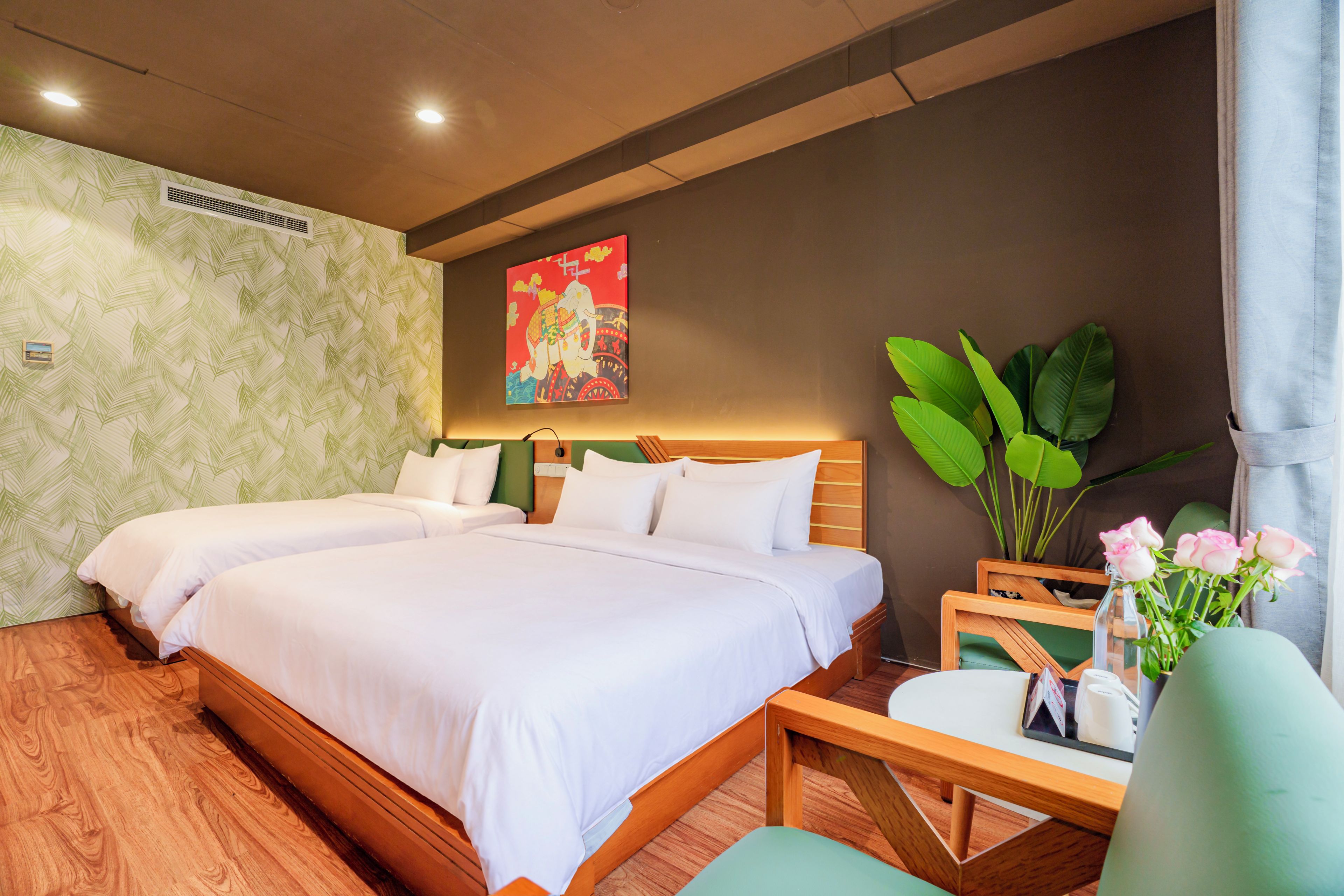 A hotel room with two beds, a desk, a chair, a large potted plant, and a wall with a tropical leaf pattern.