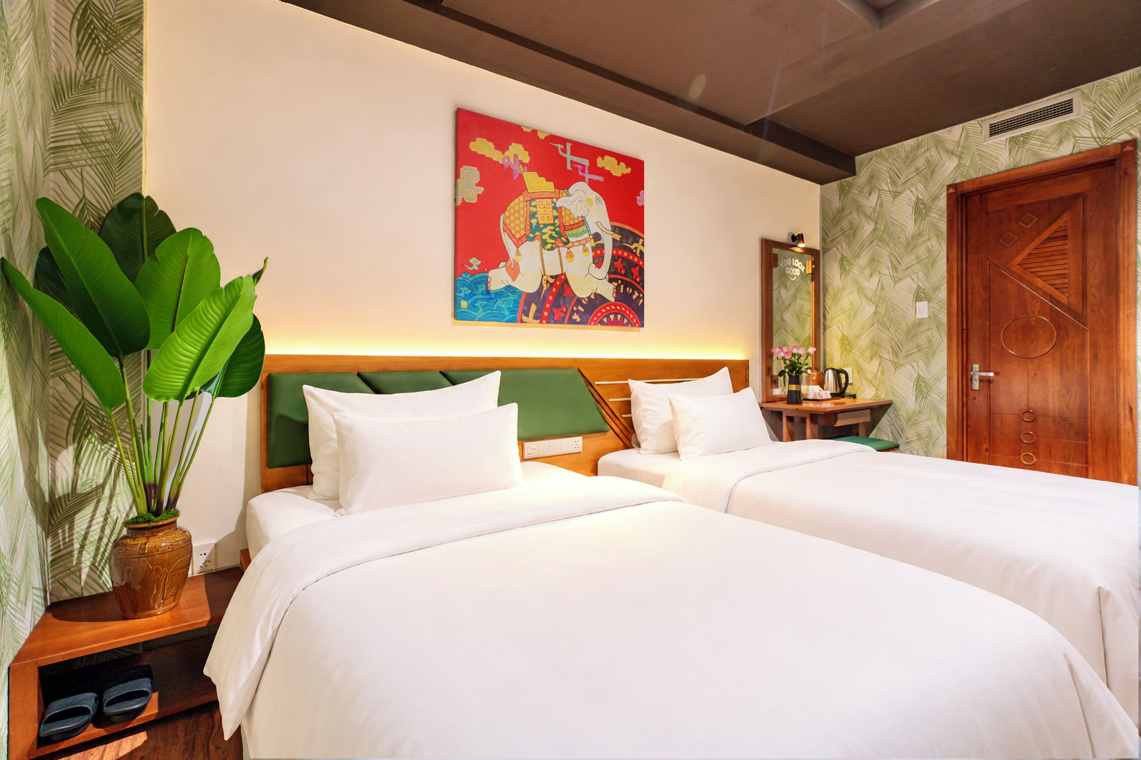Une chambre d’hôtel avec deux lits, une grande plante, un tableau et une porte en bois.