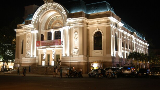 A grand, illuminated building with classical architecture, featuring a prominent entrance and steps leading up to it. - The Noble Swan Saigon (Ho Chi Minh City)