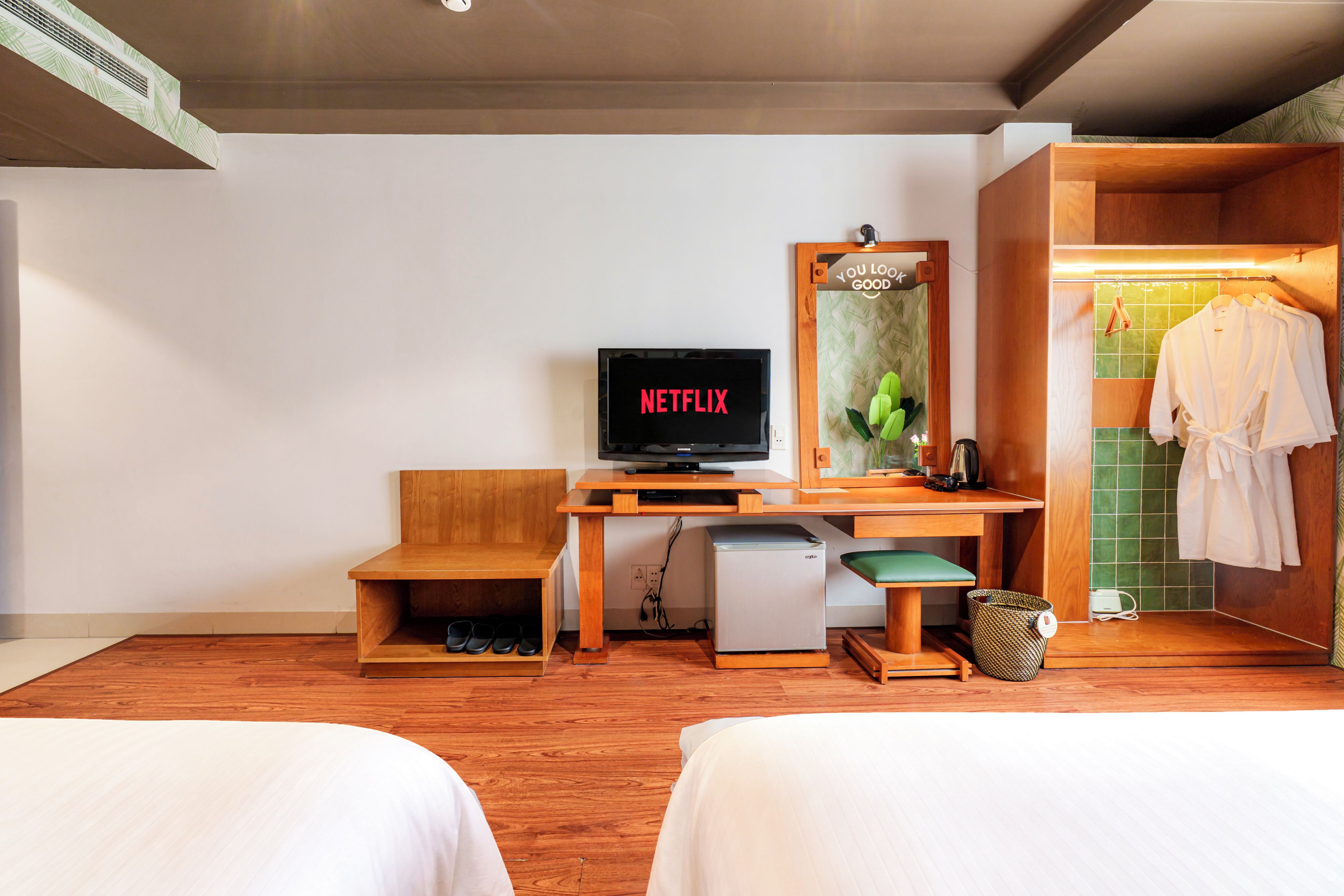 Ein Hotelzimmer mit einem Holztisch, einem Fernseher der 'NETFLIX' anzeigt, einer kleinen Kühlbox, einem Spiegel und einem Kleiderschrank mit einem Handtuch.