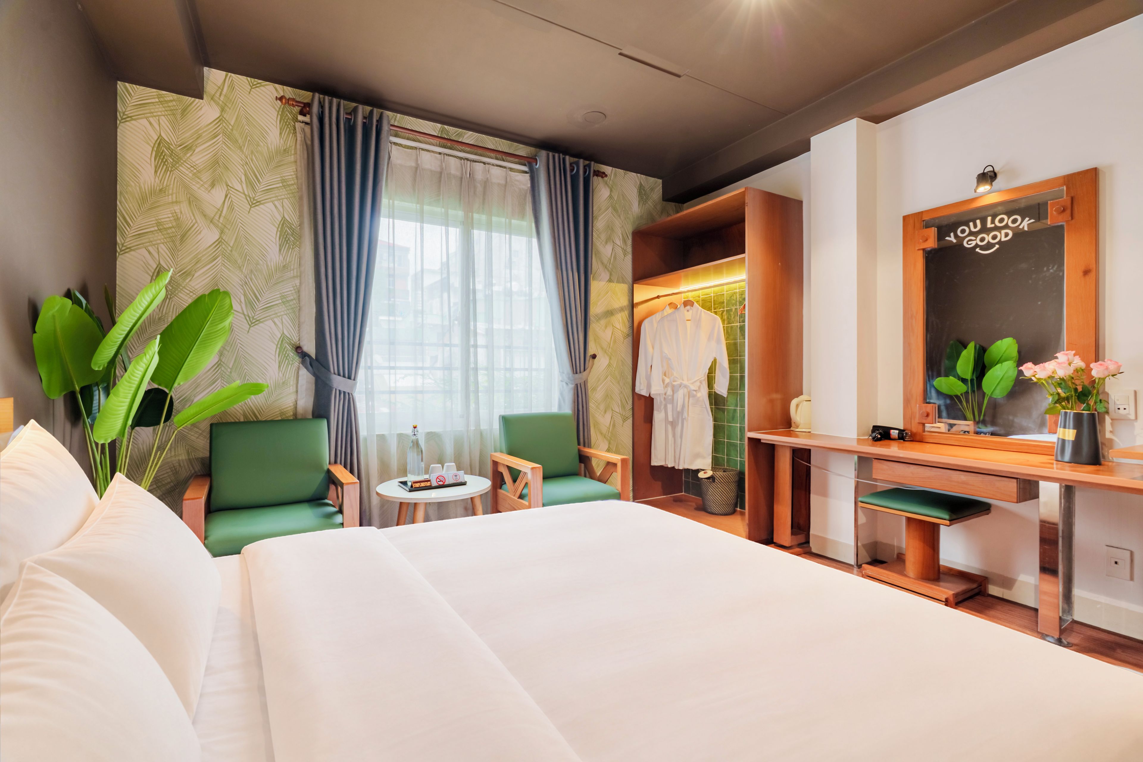 A hotel room with a bed, two chairs, a small table, a vanity with a mirror, and a view of the outside through a window.
