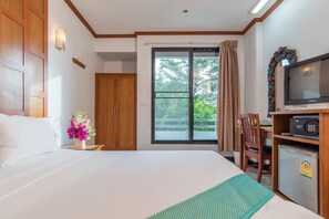 Une chambre d’hôtel comprenant un lit, un bureau avec une télévision, un réfrigérateur, une armoire en bois, une fenêtre donnant sur un paysage verdoyant et une petite table agrémentée d’un vase de fleurs.