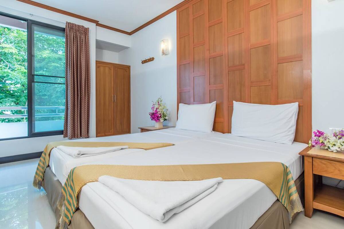 A hotel room with two beds, wooden paneling on the walls, a large window with curtains, and a nightstand with a vase of flowers.