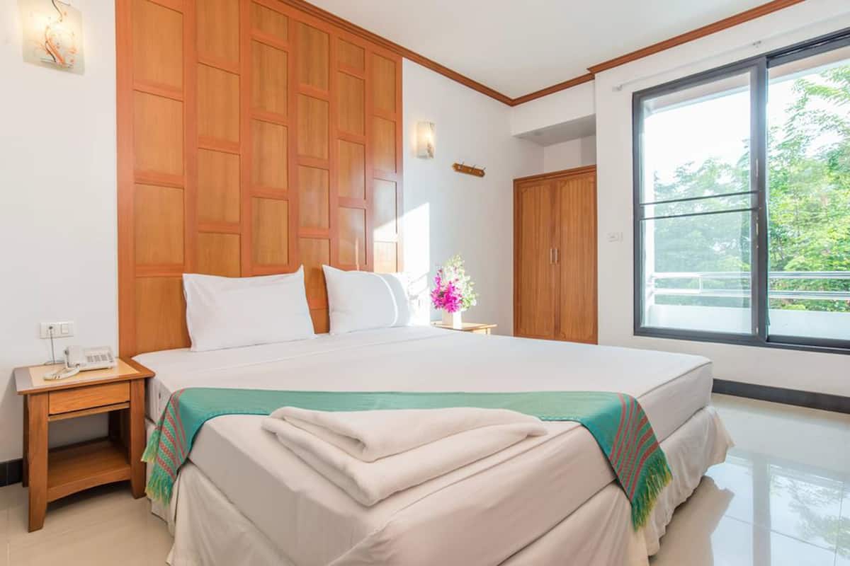 A hotel room with a large bed, wooden paneling on the walls, a nightstand with a phone, and a window with a view of greenery.