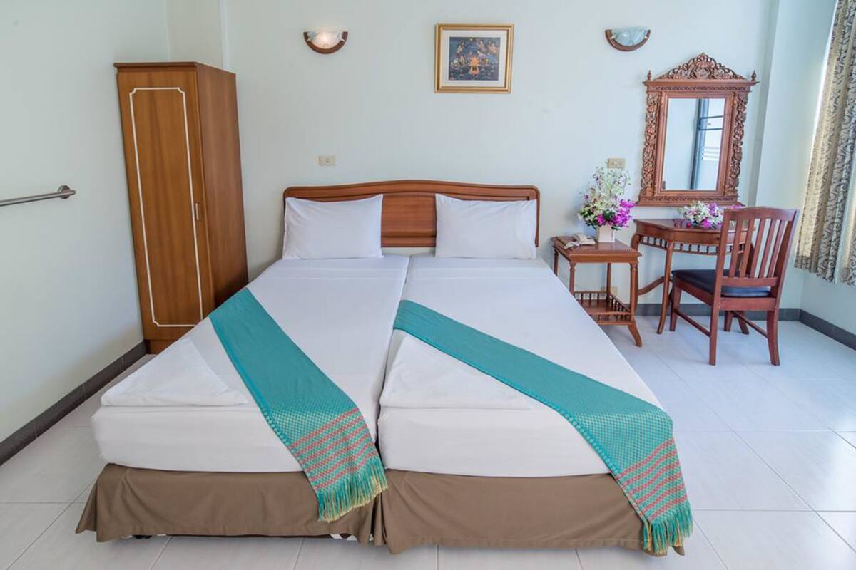 A double bed with a teal and brown blanket, a wooden wardrobe, a desk with a chair, a mirror, and a framed picture on the wall.