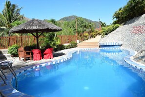 Una piscina con una estructura de techo de paja, sillas rojas y una pared de piedra.