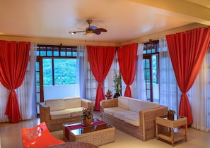 A room with wicker furniture, a ceiling fan, and large windows with red curtains.
