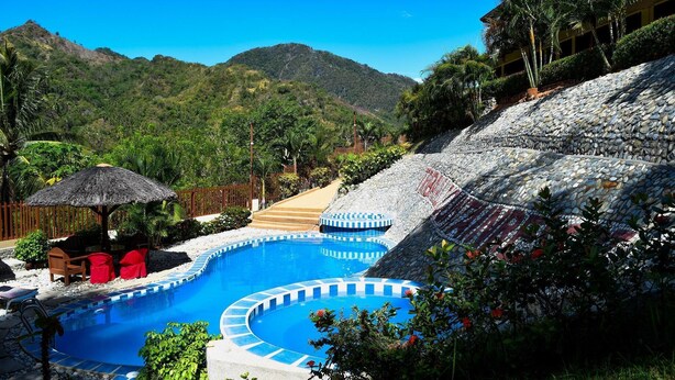 A swimming pool with a stone wall and a thatched umbrella, surrounded by greenery and a mountainous backdrop.