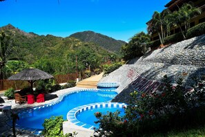 A swimming pool with a stone wall and a thatched umbrella, surrounded by greenery and a mountainous backdrop. - Tribal Hills Mountain Resort (Puerto Galera)