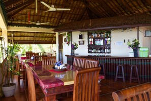 Un restaurante con mesas y sillas de madera, una zona de bar con botellas y un techo de paja.