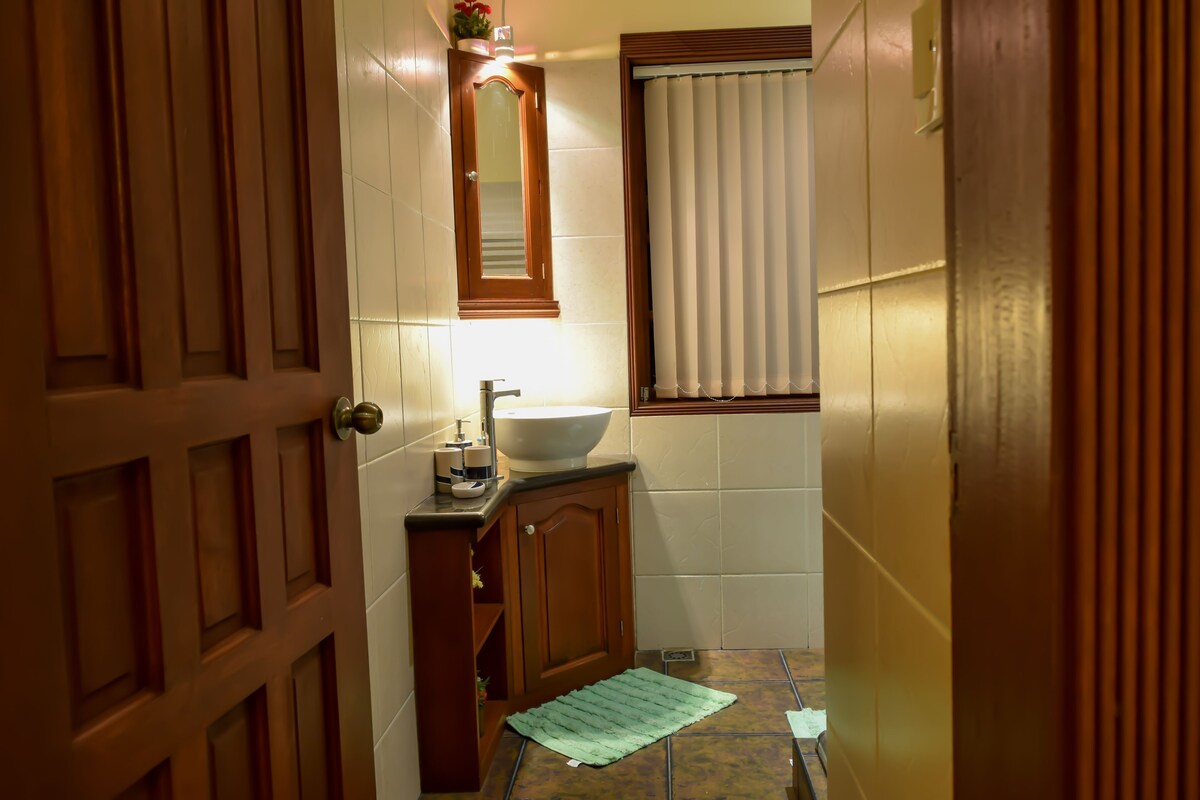 Un baño con puerta de madera, un lavabo con espejo arriba y una ventana con persianas.