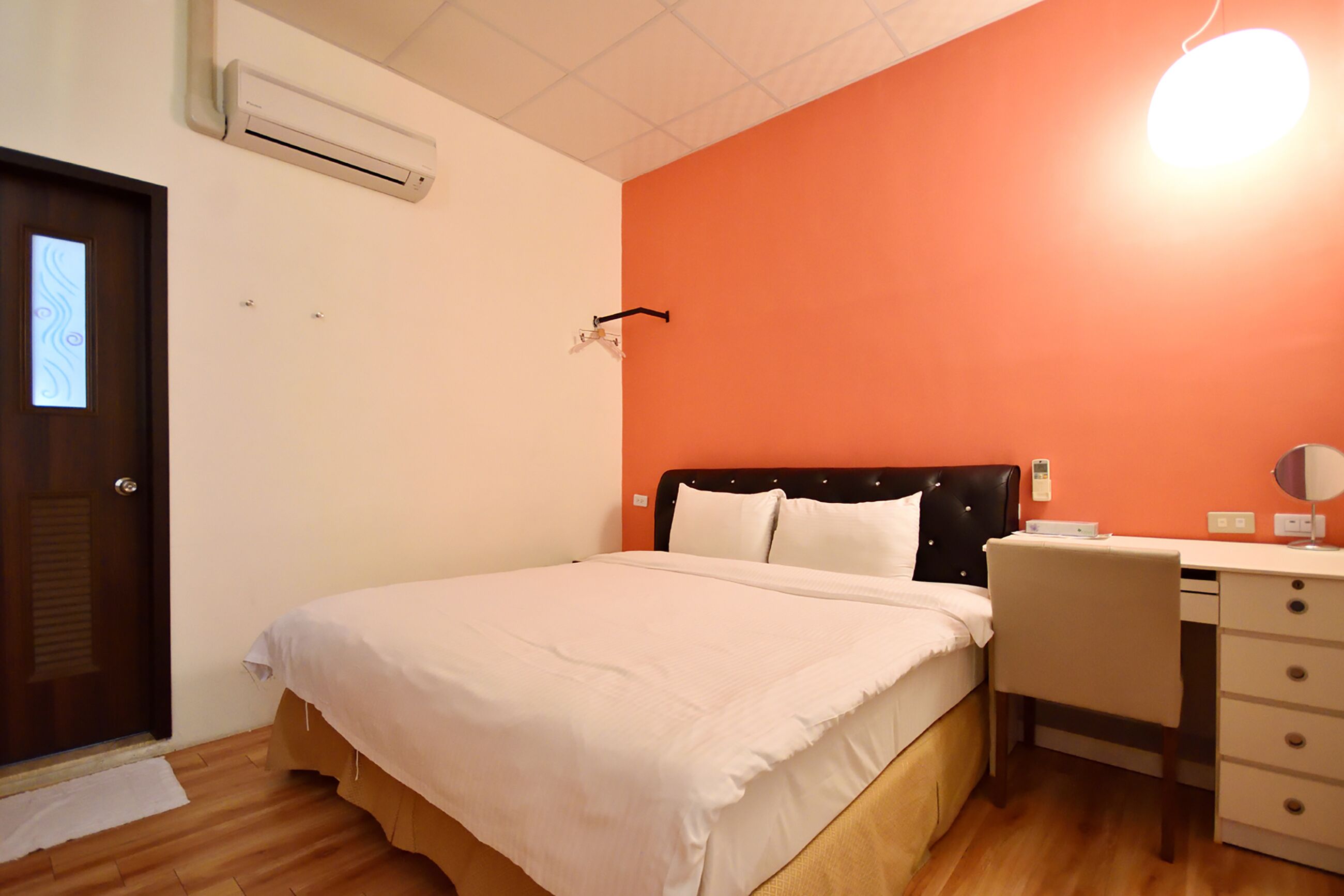 A bedroom with a bed, a desk, and a chair. There is an air conditioning unit on the wall and a door with a light panel.