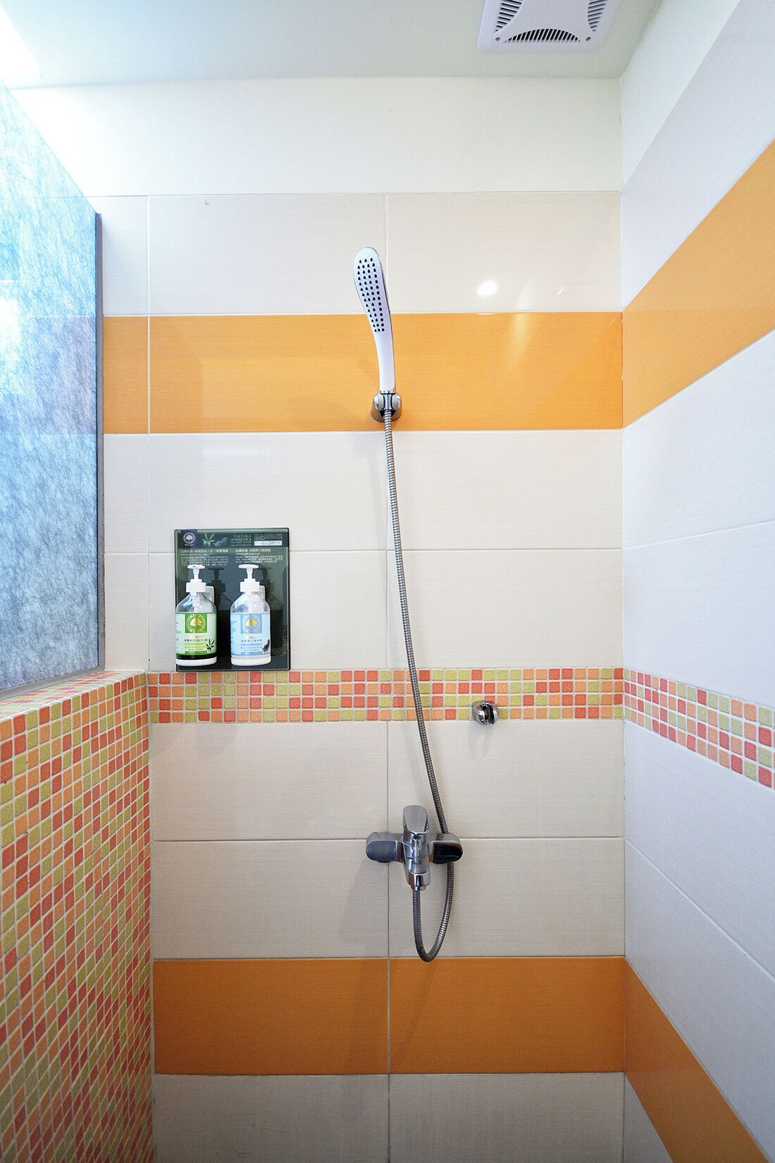 Zona de ducha con cabezal de ducha manual, grifo empotrado en la pared y dos botellas de champú y acondicionador sobre una estantería.