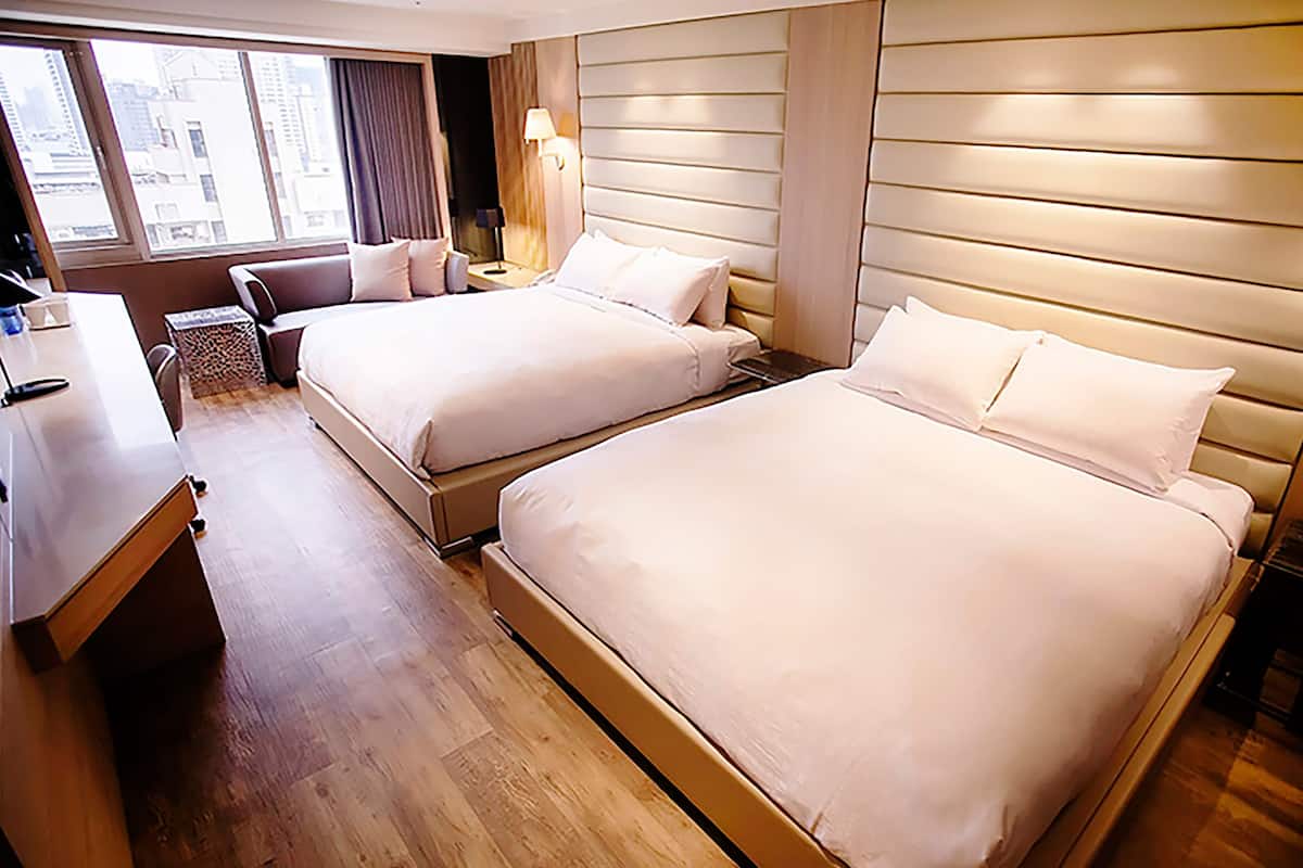 Ein Hotelzimmer mit zwei Betten, einem Schreibtisch und einem Fenster mit Blick auf Gebäude.
