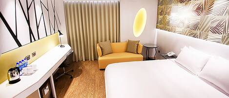 A hotel room with a bed, a desk with a lamp, a sofa, a small table, and a wall with a leaf pattern.